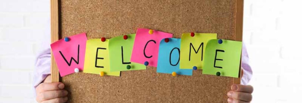 Woman holding corkboard with word Welcome near white brick wall, closeup
